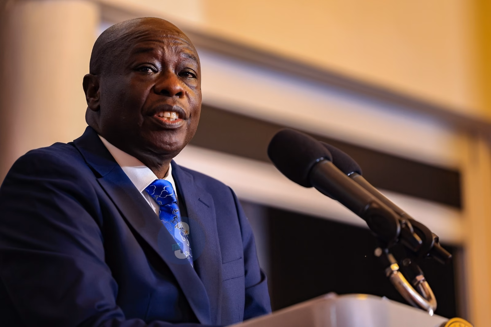 Rigathi Gachagua speaking at a podium in a dark blue suit and a patterned blue tie.