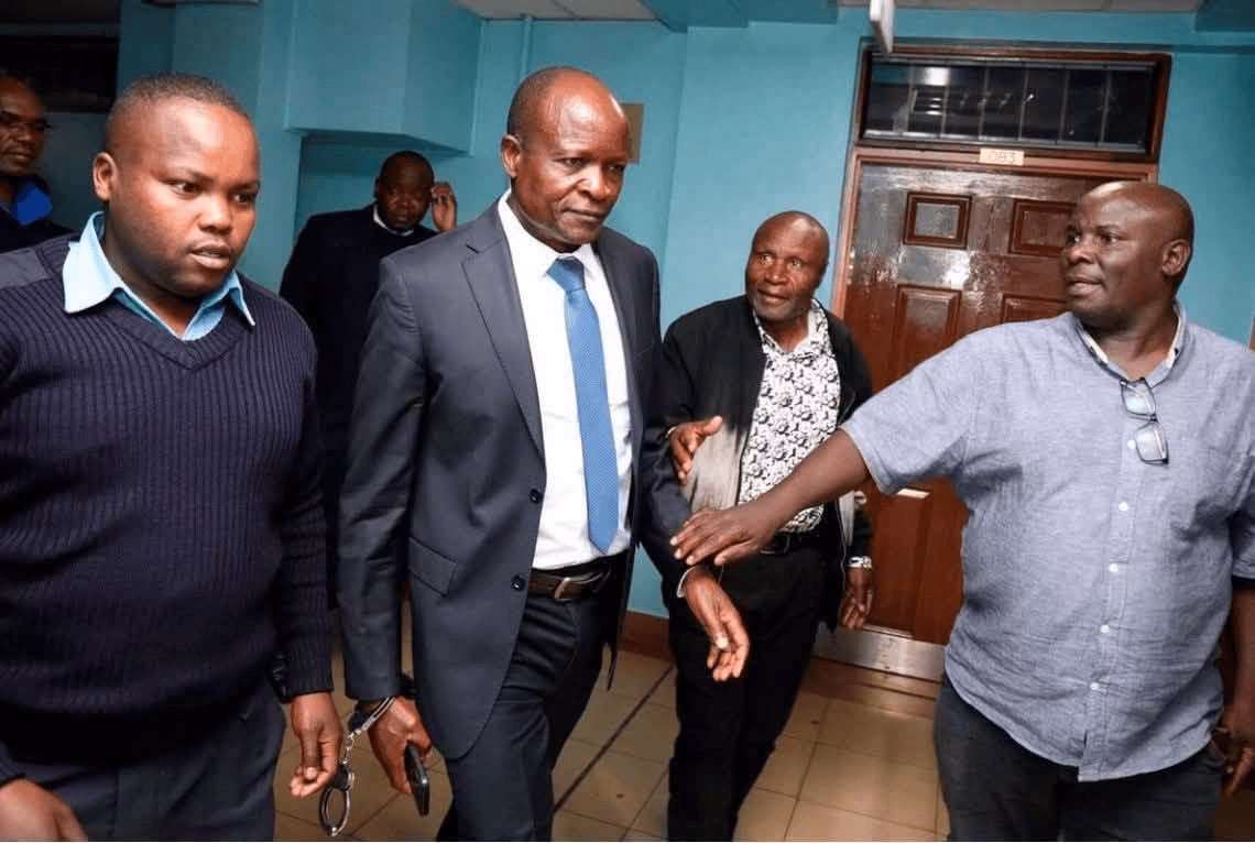 Former Migori Governor Okoth Obado wearing a suit and handcuffs being escorted inside a courtroom corridors.