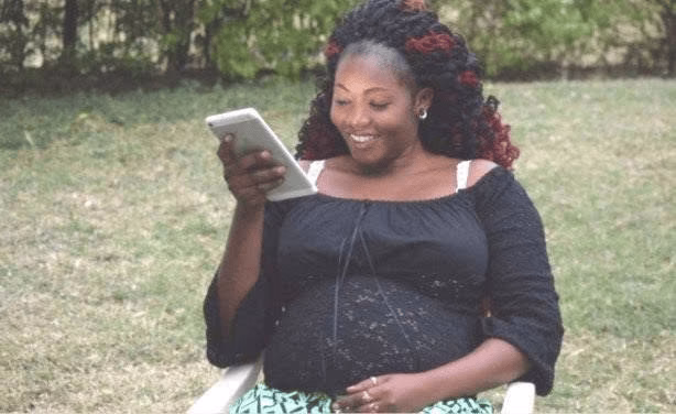 Pregnant Sharon Otieno sitting outdoors on a chair holding a smartphone and smiling.