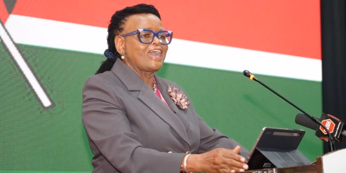 Chief Justice Martha Koome speaking at a podium with a microphone, wearing a grey suit and blue glasses against a Kenyan flag background.