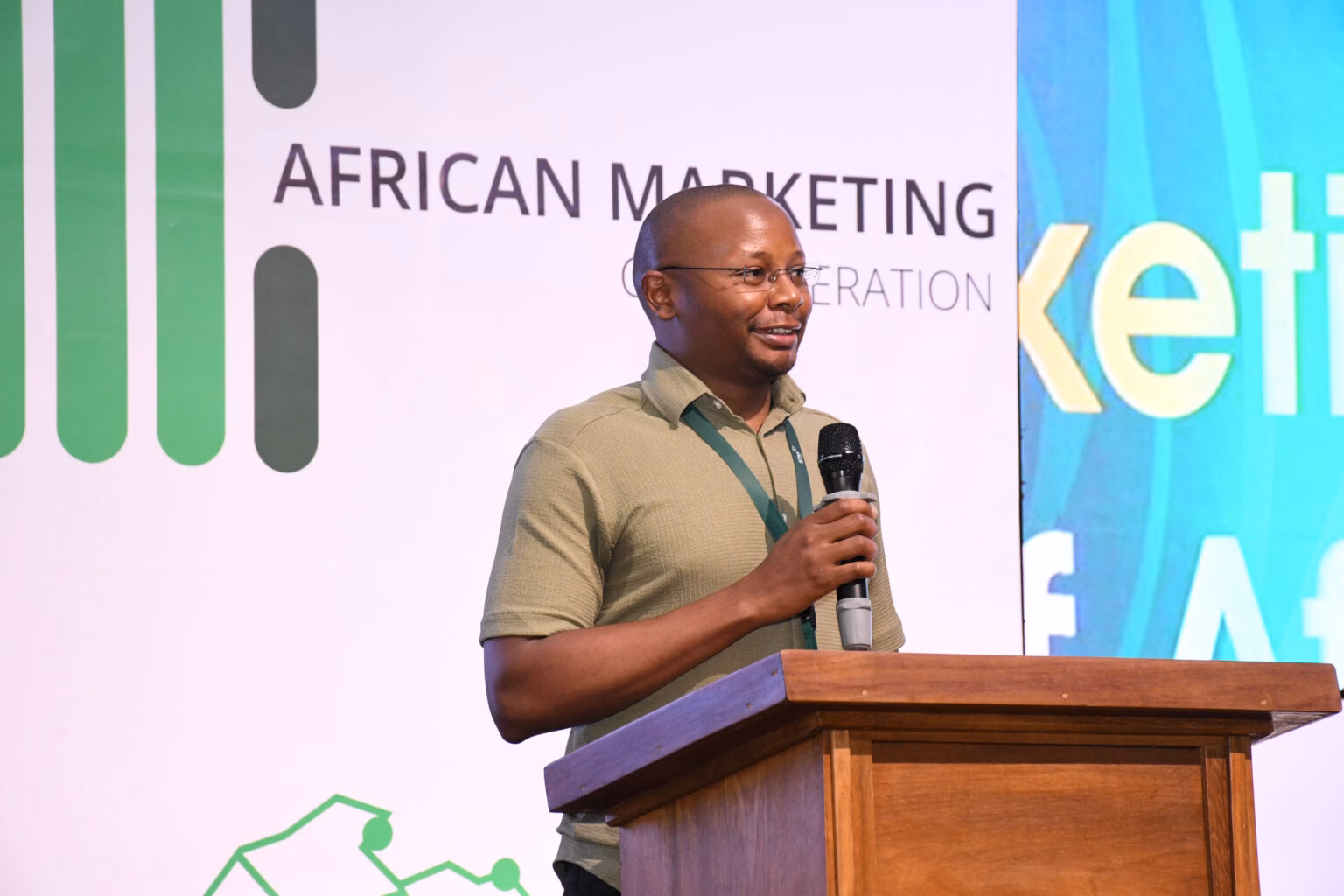 Dr. Adrian Kamotho holding a microphone and speaking at a wooden podium during a conference.