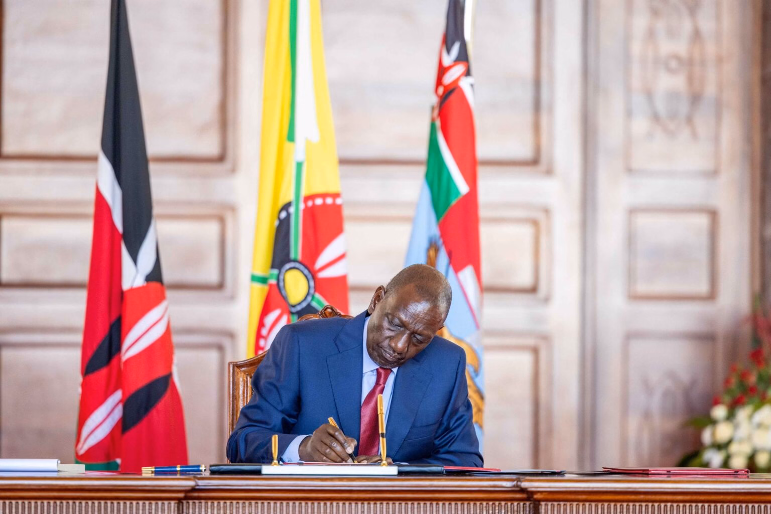 Kenyan President William Ruto sitting at a wooden desk signing official executive documents with the Kenyan flag and Presidential standard in the background.