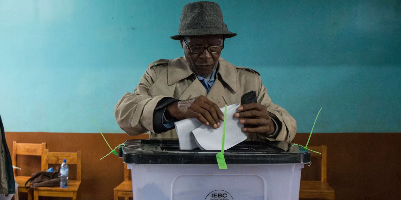 An elderly man wearing a beige trench coat and hat folding his ballot paper over a clear IEBC ballot box